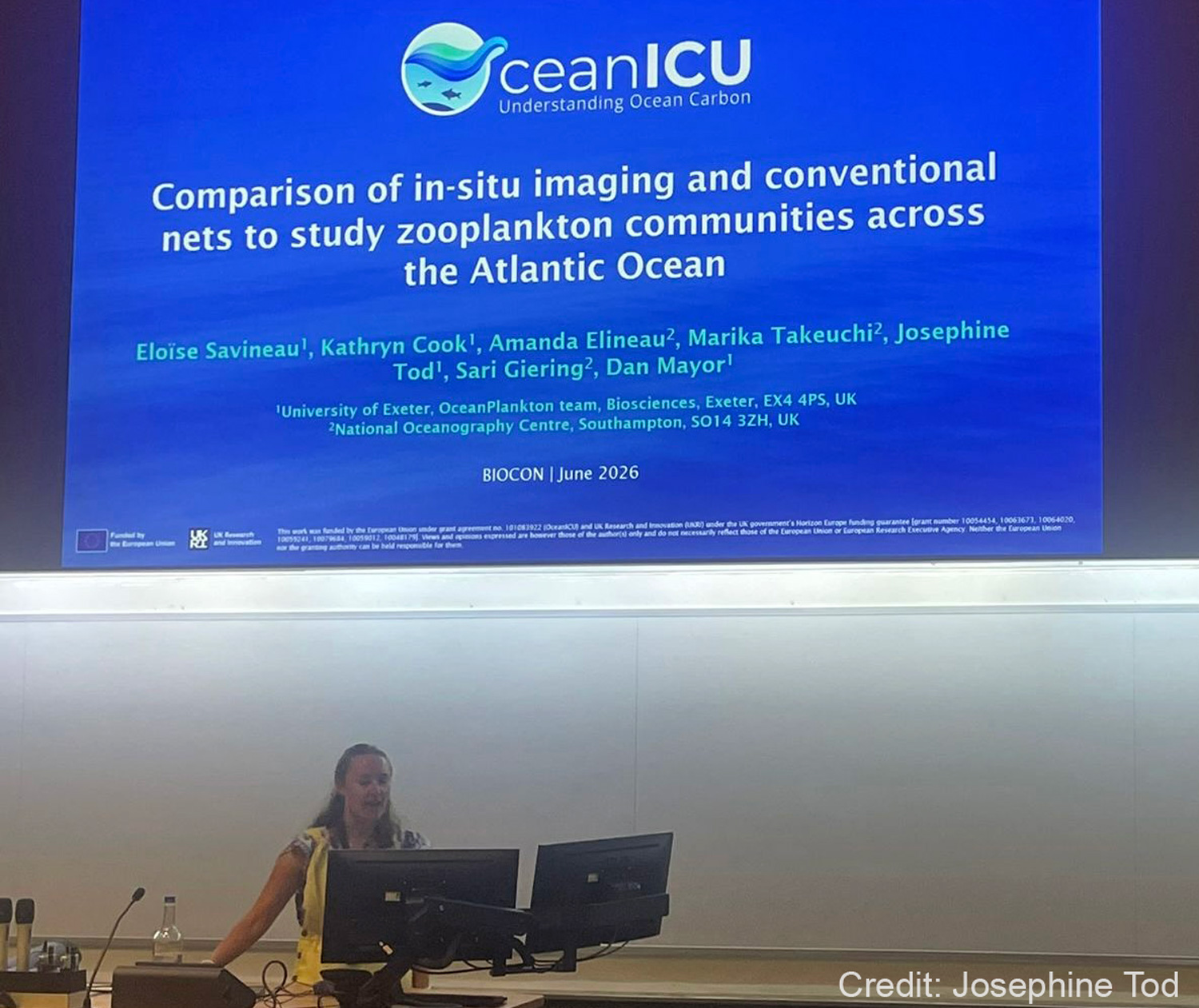 A woman giving a presentation on stage, with a big screen behind her.
