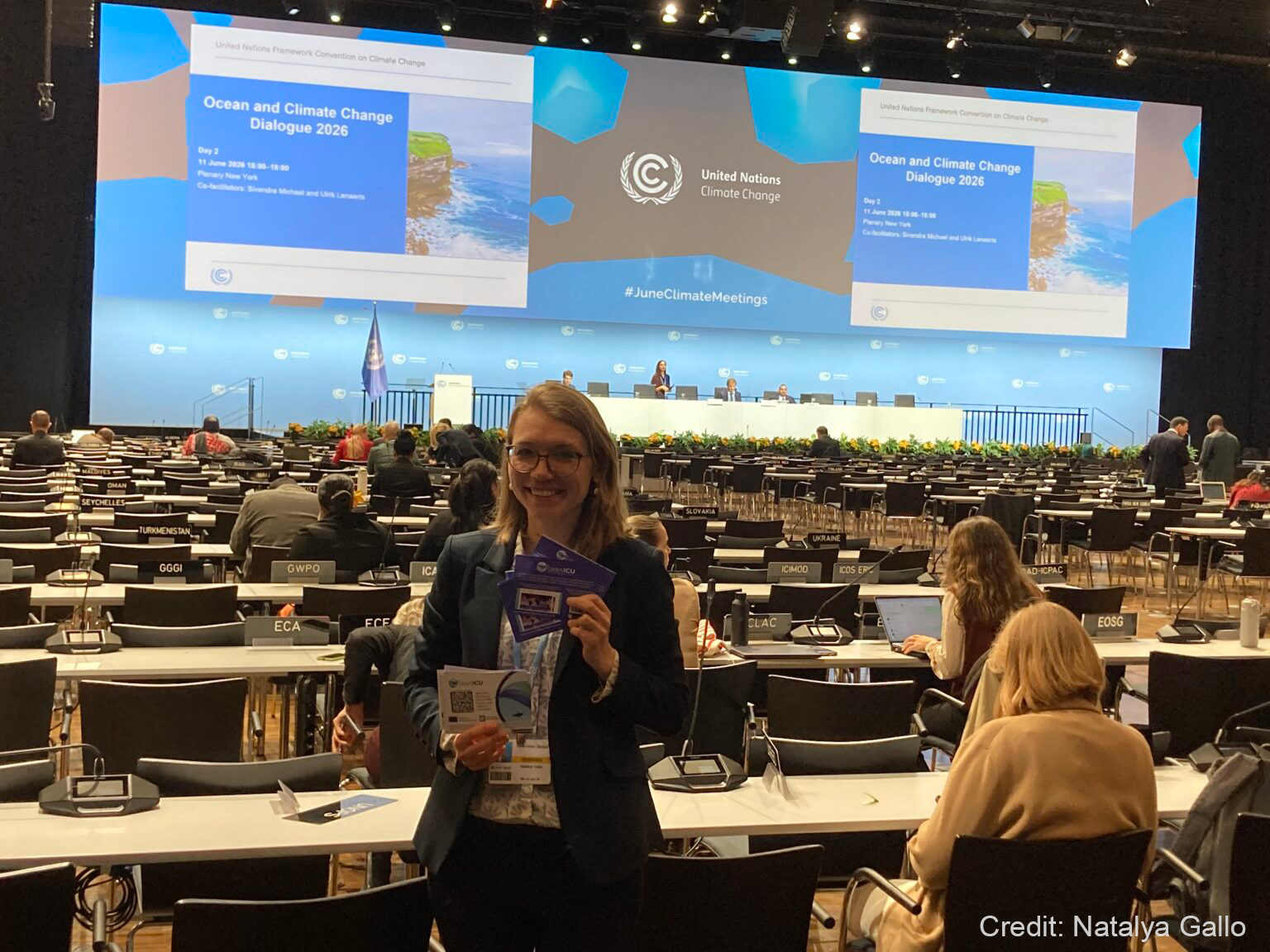 A woman with glasses smiling at the camera, holding OceanICU flyers. In the background are many desks and chairs facing a podium and a large screen showing 'United Nations Climate Change: Ocean and Climate Change Dialogue 2026'