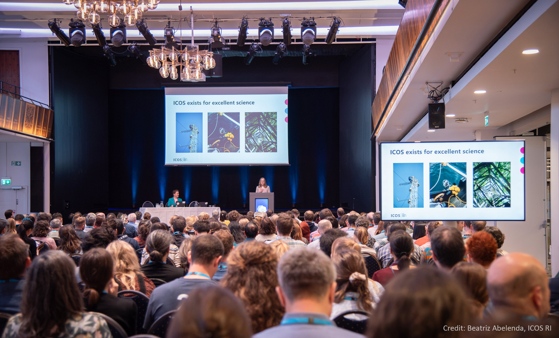 A photo from ICOS Science Conference taken standing from behind the audience, showing a crowd sitting at a conference looking at the stage with 2 screens and a speaker giving a presentation.