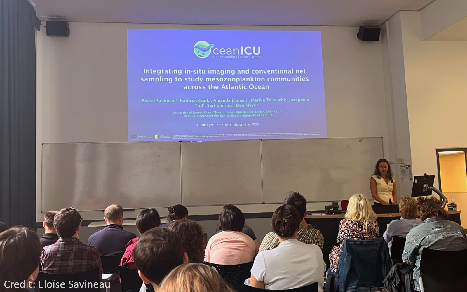 A photo from Challenger Society for Marine Science Conference taken standing from behind the audience, showing a crowd sitting at a conference looking at the stage with a woman giving a presentation.